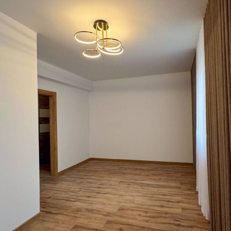 Empty living room with wood flooring, white walls, and brown curtains along the right; a circular ceiling light hangs overhead.