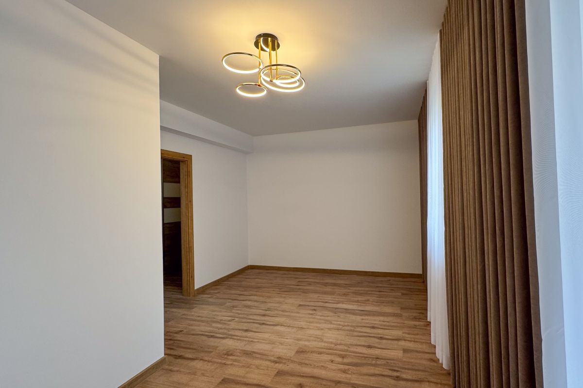 Empty living room with wood flooring, white walls, and brown curtains along the right; a circular ceiling light hangs overhead.