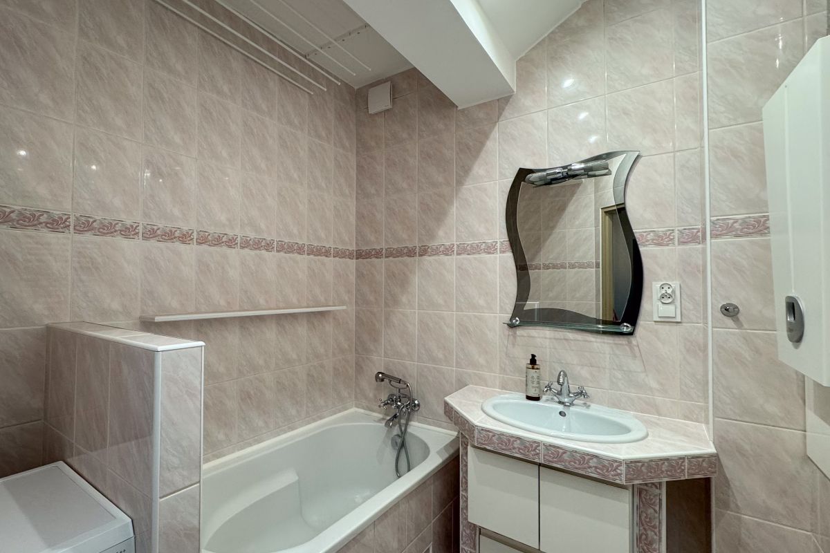 Pink-tiled bathroom with a white bathtub, oval sink vanity, decorative curved mirror, and a washing machine in the corner.