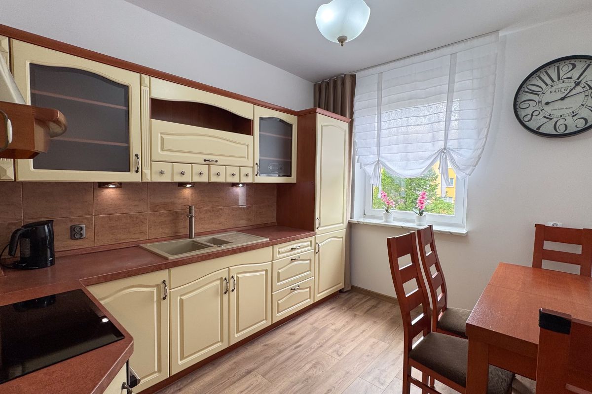 Cozy kitchen with cream cabinets, brown countertops, double sink, and a wooden dining table by a window with sheer curtains.