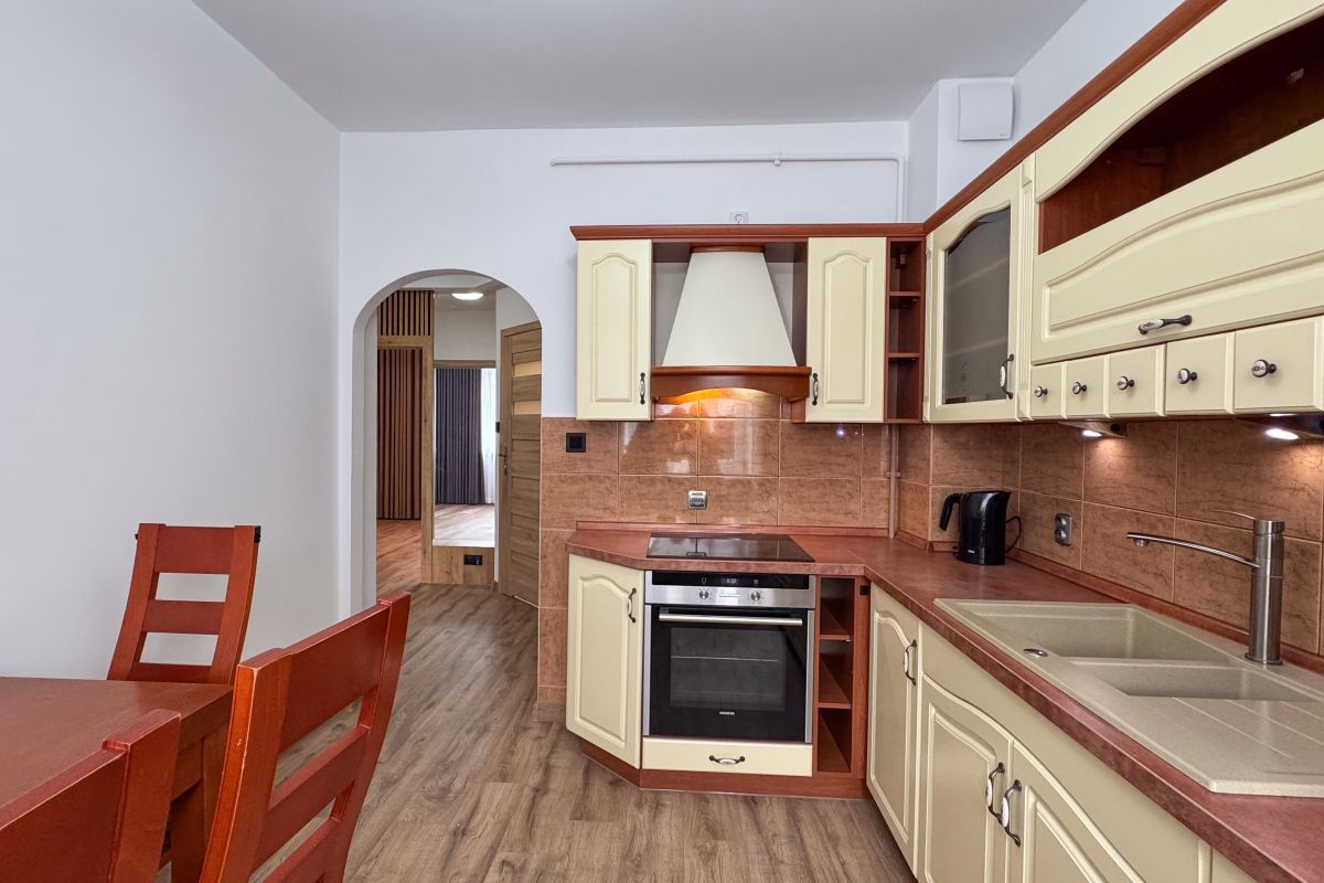 Cream kitchen with brown countertops and backsplash, built-in oven and stovetop, range hood, and double sink; wooden dining table in foreground.