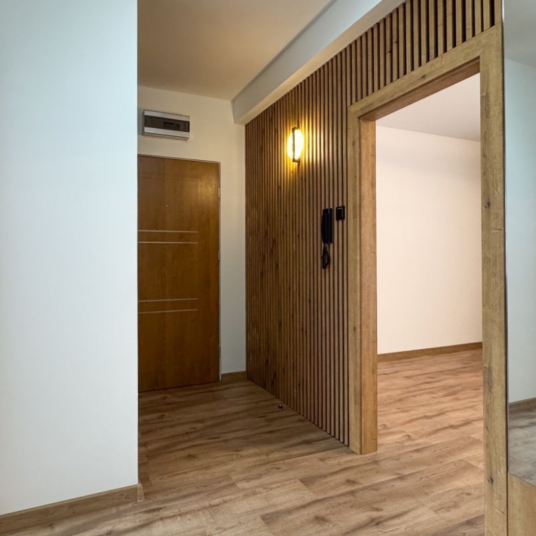 Bright hallway with a vertical wooden slat partition, a warm wall light, and wooden doors; light-colored walls and wood flooring create a modern entry area.