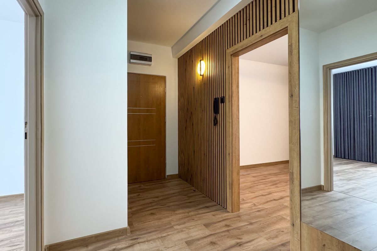Bright hallway with a vertical wooden slat partition, a warm wall light, and wooden doors; light-colored walls and wood flooring create a modern entry area.