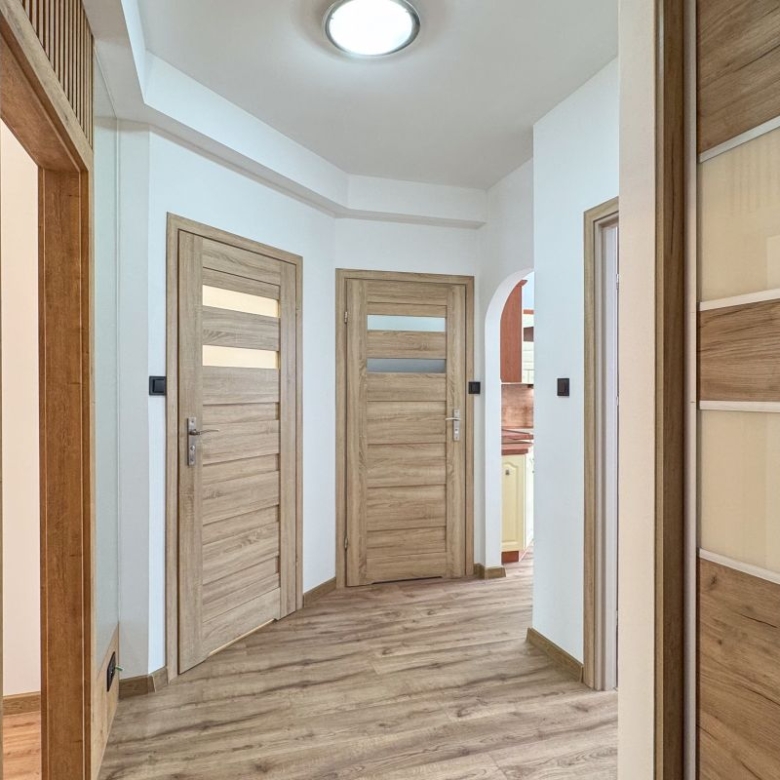 Bright hallway with three wooden doors, light wood flooring, and a wall-mounted intercom on the left.