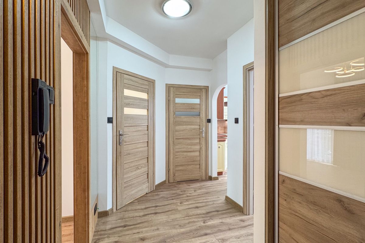 Bright hallway with three wooden doors, light wood flooring, and a wall-mounted intercom on the left.