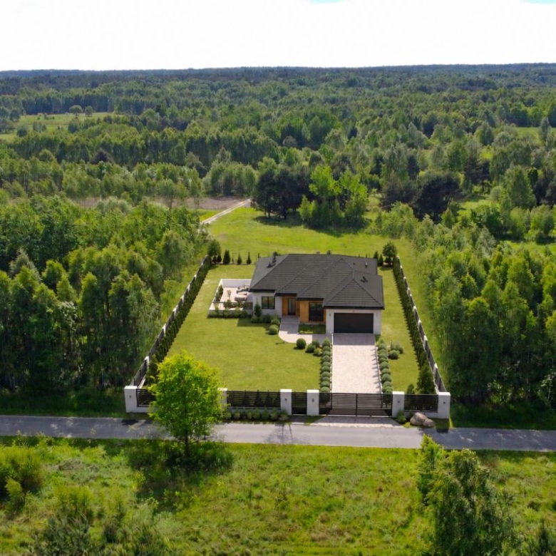 Aerial view of a modern single-family home on a large grassy lot, surrounded by trees with a fence and gate at the front.