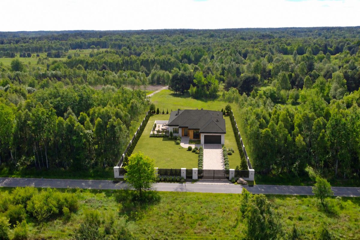 Aerial view of a modern single-family home on a large grassy lot, surrounded by trees with a fence and gate at the front.