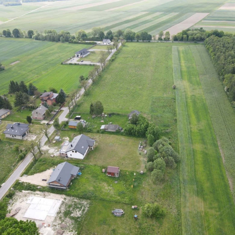 Aerial view of a rural village with houses along a road, surrounded by large green fields and a tree-lined boundary.