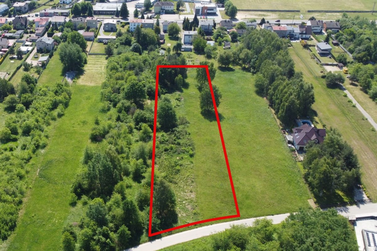 Aerial view of a vacant lot outlined in red in a green residential area, with trees along the edges and nearby houses.