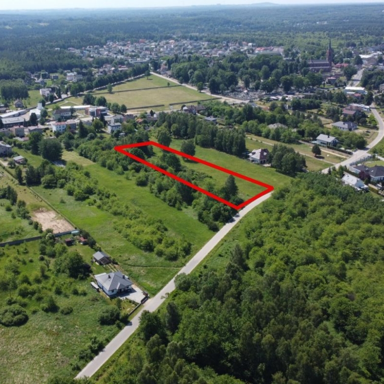 Aerial view of a grassy residential plot outlined in red, adjacent to a road near a tree-filled neighborhood.