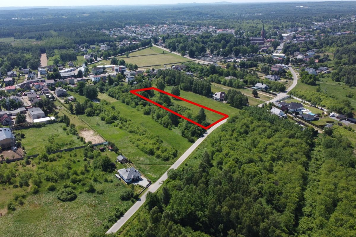 Aerial view of a grassy residential plot outlined in red, adjacent to a road near a tree-filled neighborhood.