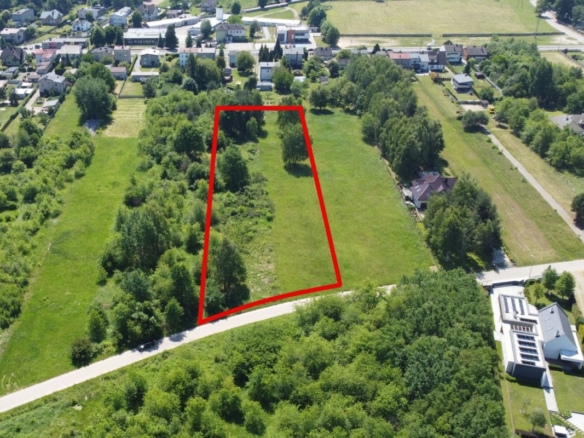 A vacant grassy lot outlined in red, bordered by trees, with a curved road along the front in a residential area in the background.