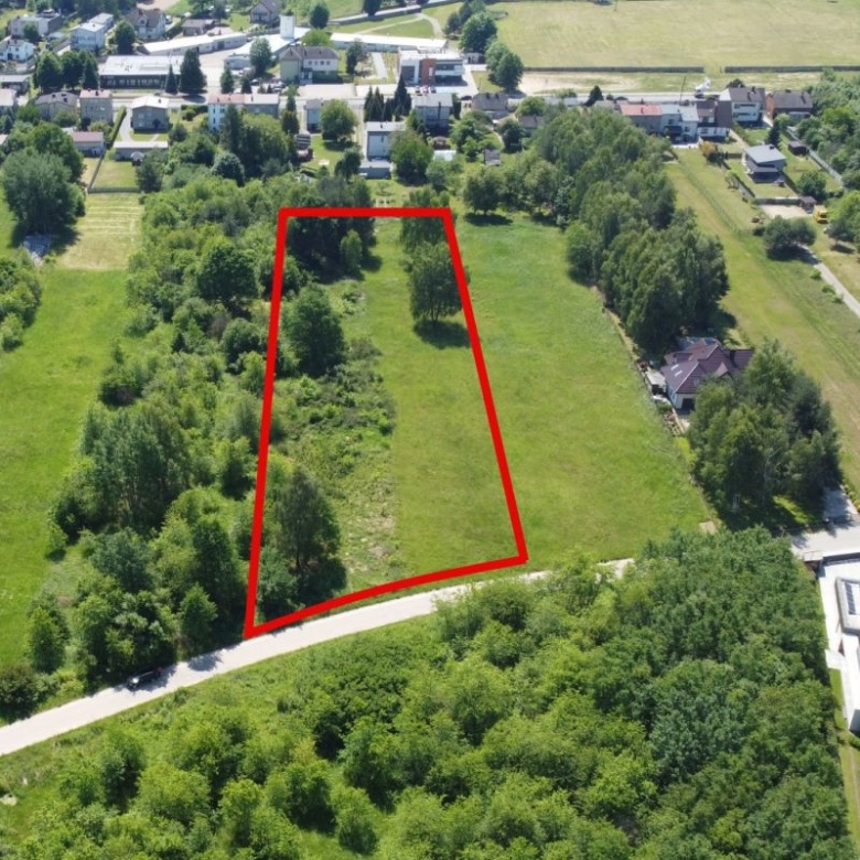 A vacant grassy lot outlined in red, bordered by trees, with a curved road along the front in a residential area in the background.