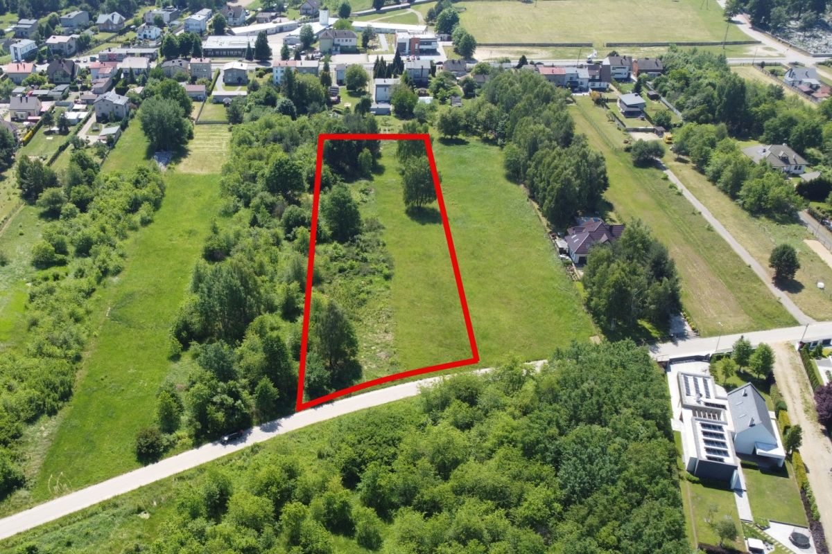 A vacant grassy lot outlined in red, bordered by trees, with a curved road along the front in a residential area in the background.