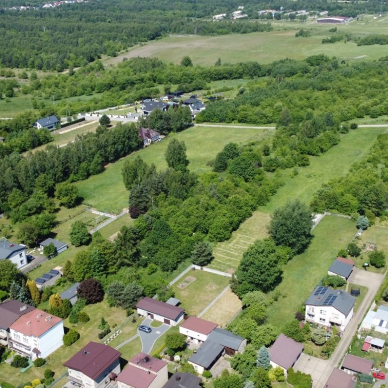 Aerial view of a suburban neighborhood with houses lining streets and large grassy lots amid many trees and open fields nearby.