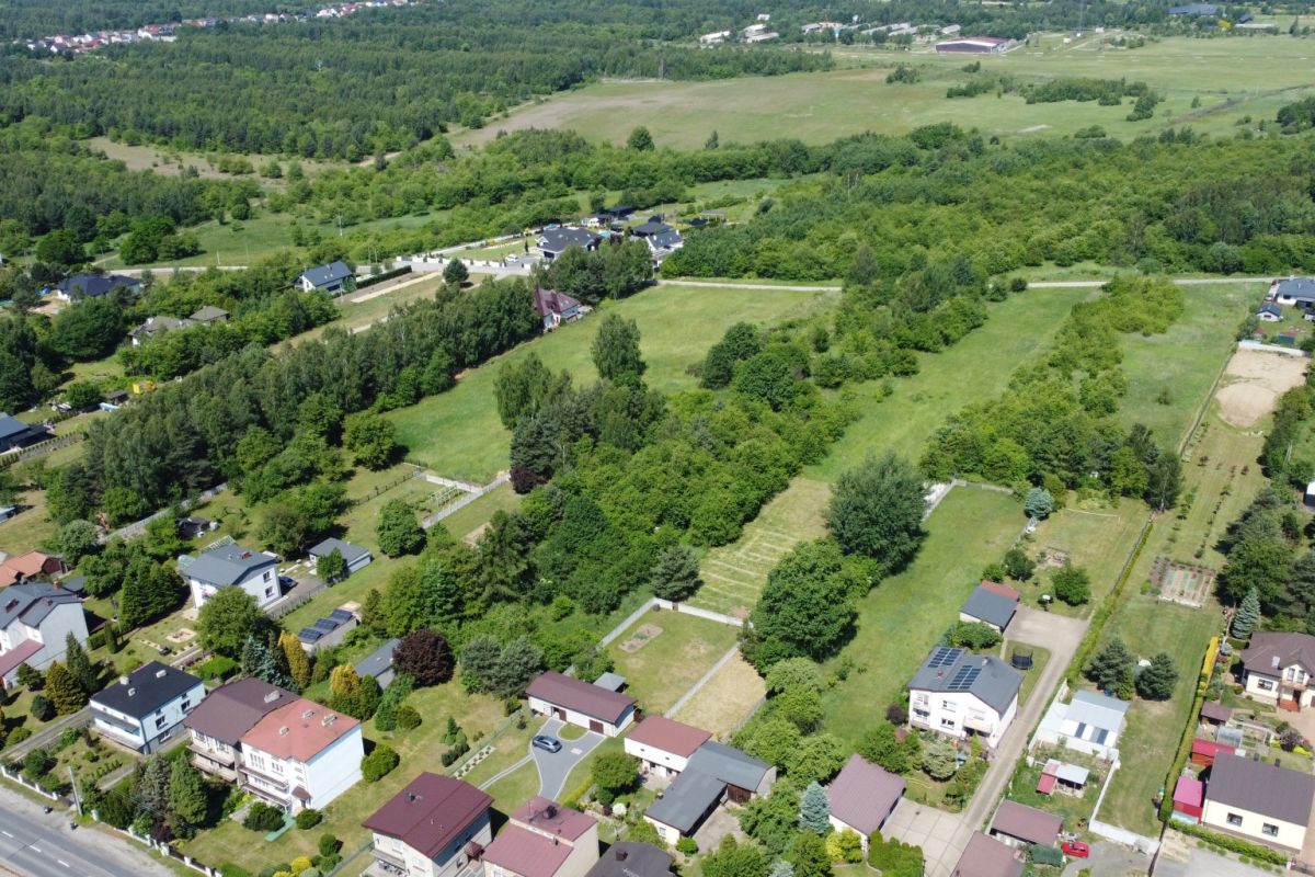 Aerial view of a suburban neighborhood with houses lining streets and large grassy lots amid many trees and open fields nearby.