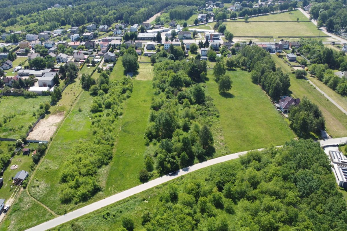 Aerial view of a suburban neighborhood adjacent to large grassy fields, with a curved road at the bottom and clusters of trees throughout.
