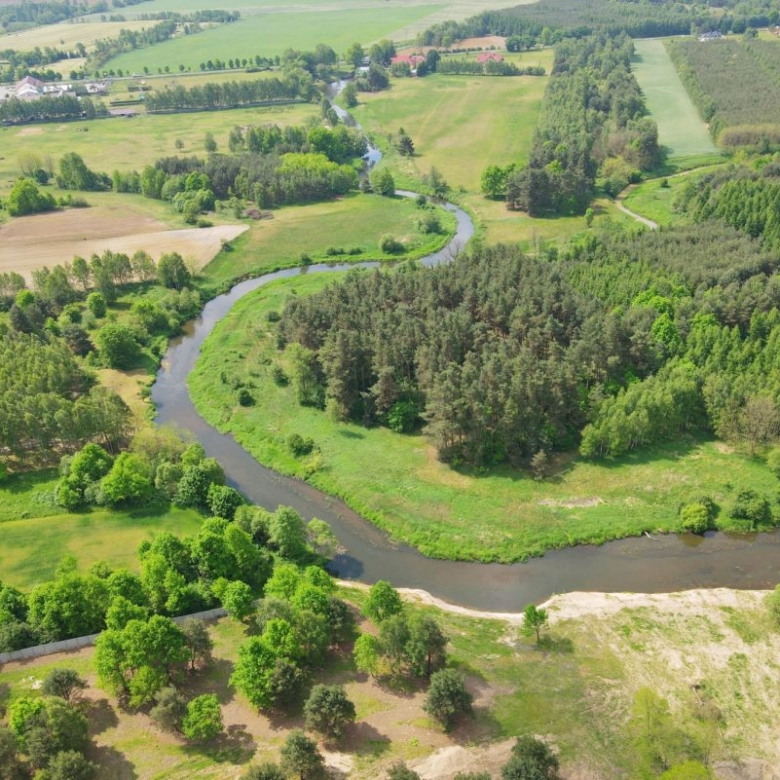 Aerial view of a winding river through green fields and forested areas with scattered farms in the distance, under a bright sky.