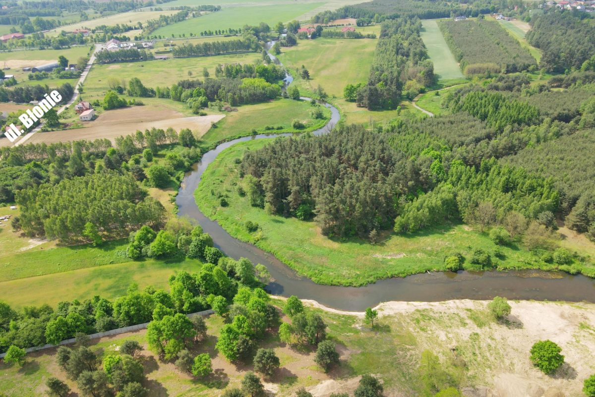 Aerial view of a winding river through green fields and forested areas with scattered farms in the distance, under a bright sky.