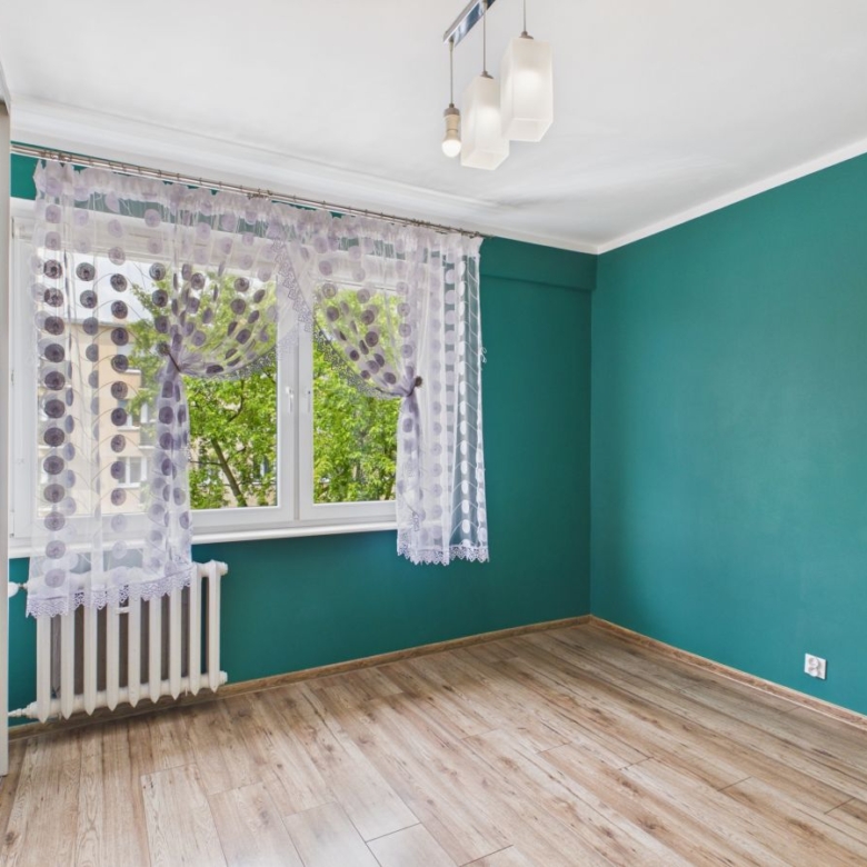 Empty room with teal walls, light wood floor, and a window with sheer lace curtains near a radiator