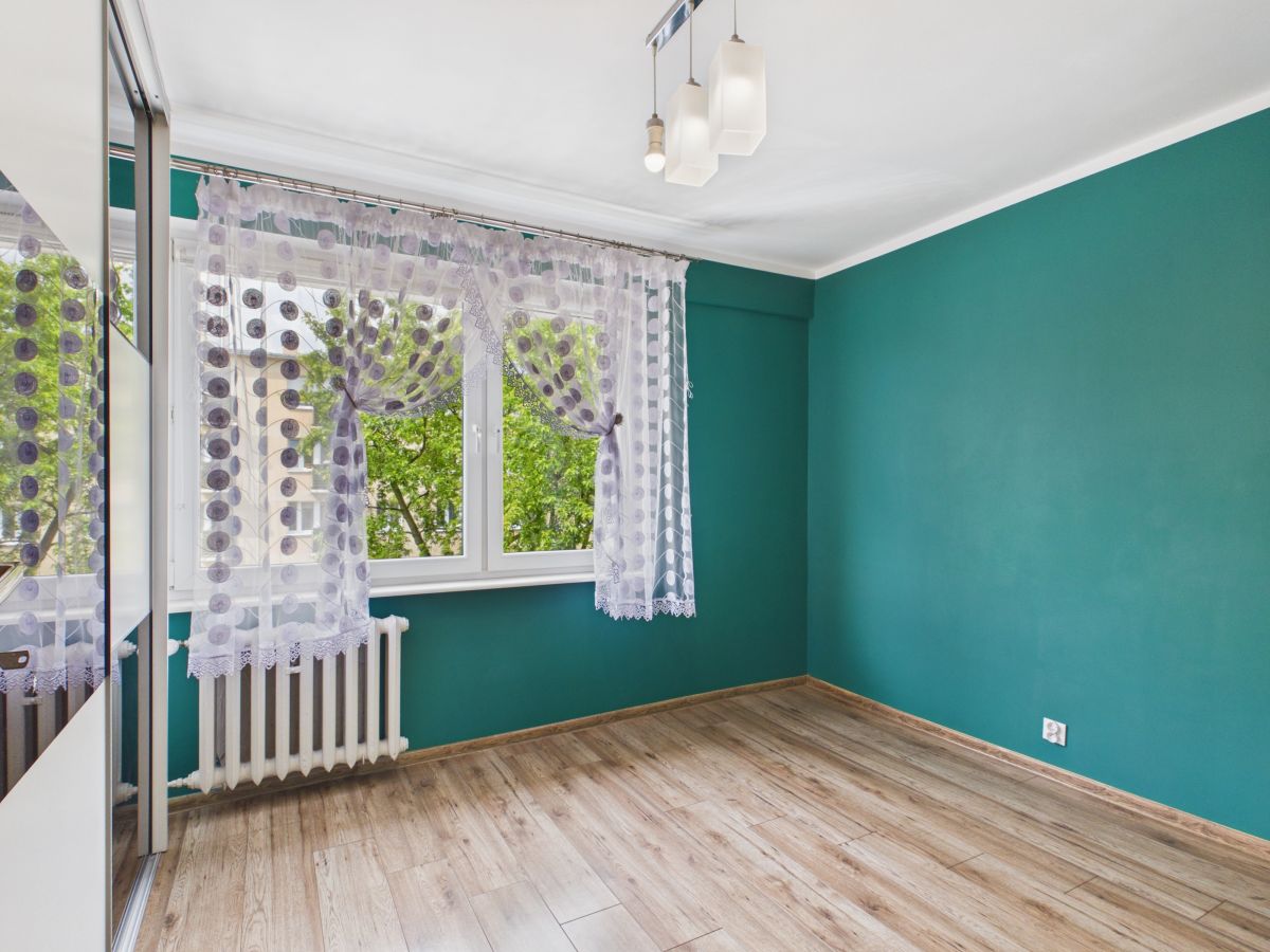 Empty room with teal walls, light wood floor, and a window with sheer lace curtains near a radiator