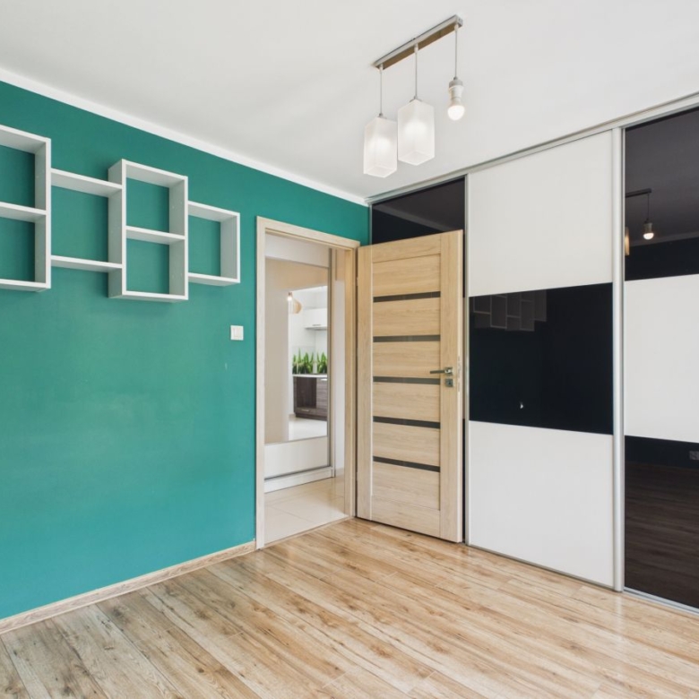 Teal accent wall with white geometric wall shelves in a modern room, open wooden door, and sliding black-and-white wardrobe panels.