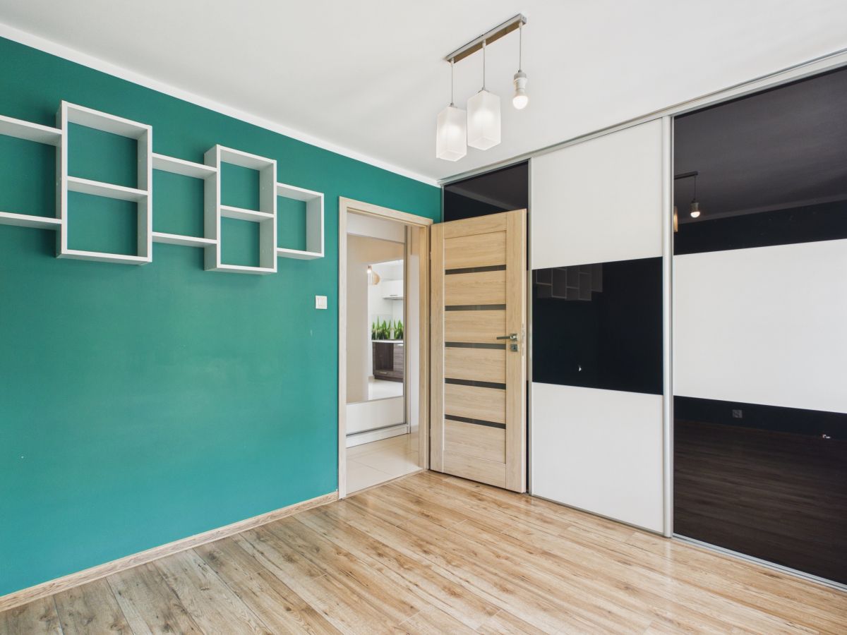 Teal accent wall with white geometric wall shelves in a modern room, open wooden door, and sliding black-and-white wardrobe panels.