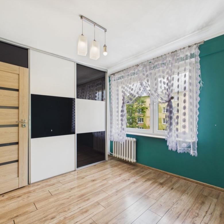 Modern bedroom corner with a wooden door, sliding wardrobe, and a large window with lace curtains on a teal wall.