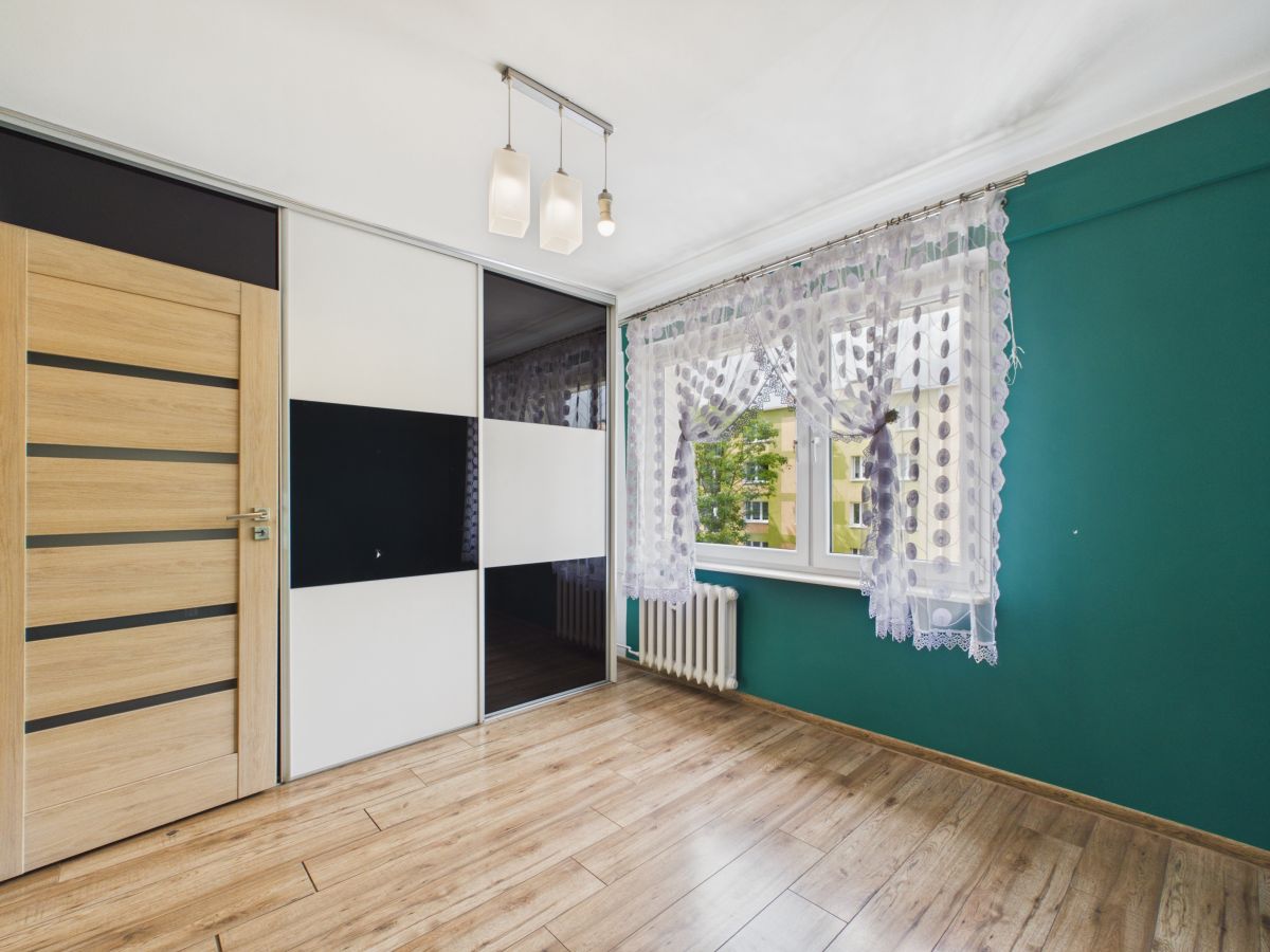 Modern bedroom corner with a wooden door, sliding wardrobe, and a large window with lace curtains on a teal wall.