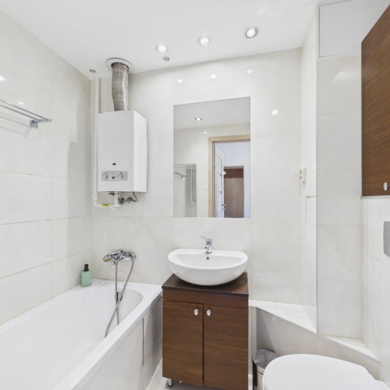 Modern white-tiled bathroom with a bathtub on the left, a wall-mounted boiler, a round vessel sink on a wooden vanity, a rectangular mirror, and a wall-mounted toilet with concealed cistern.