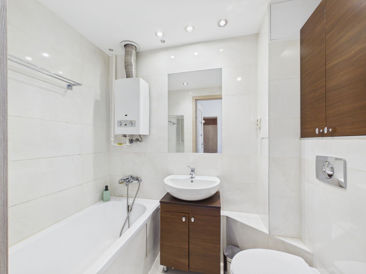 Modern white-tiled bathroom with a bathtub on the left, a wall-mounted boiler, a round vessel sink on a wooden vanity, a rectangular mirror, and a wall-mounted toilet with concealed cistern.