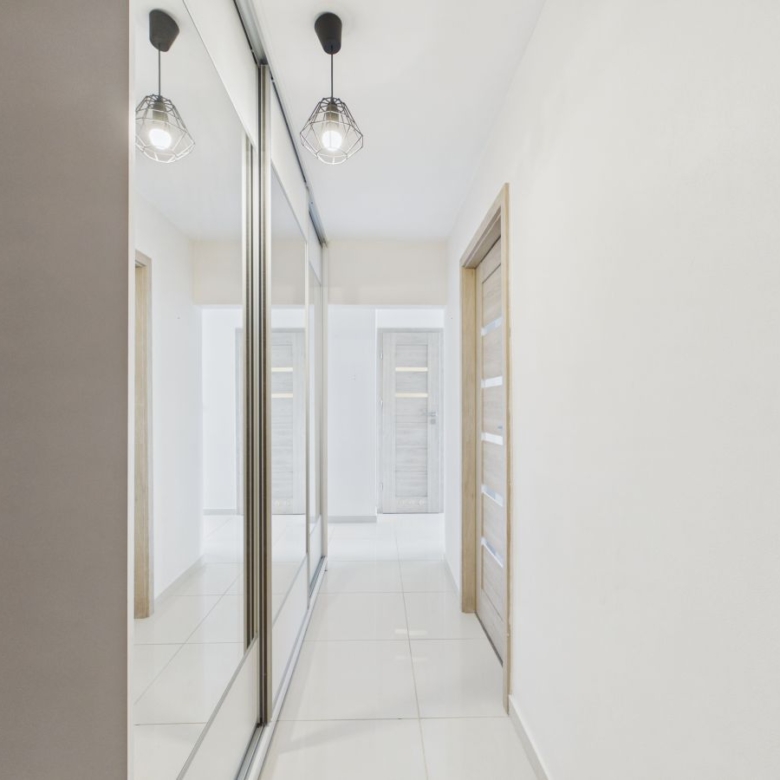 Bright, minimalist hallway with glossy white tile floor, mirrored closets on the left, and light wood doors on the right.