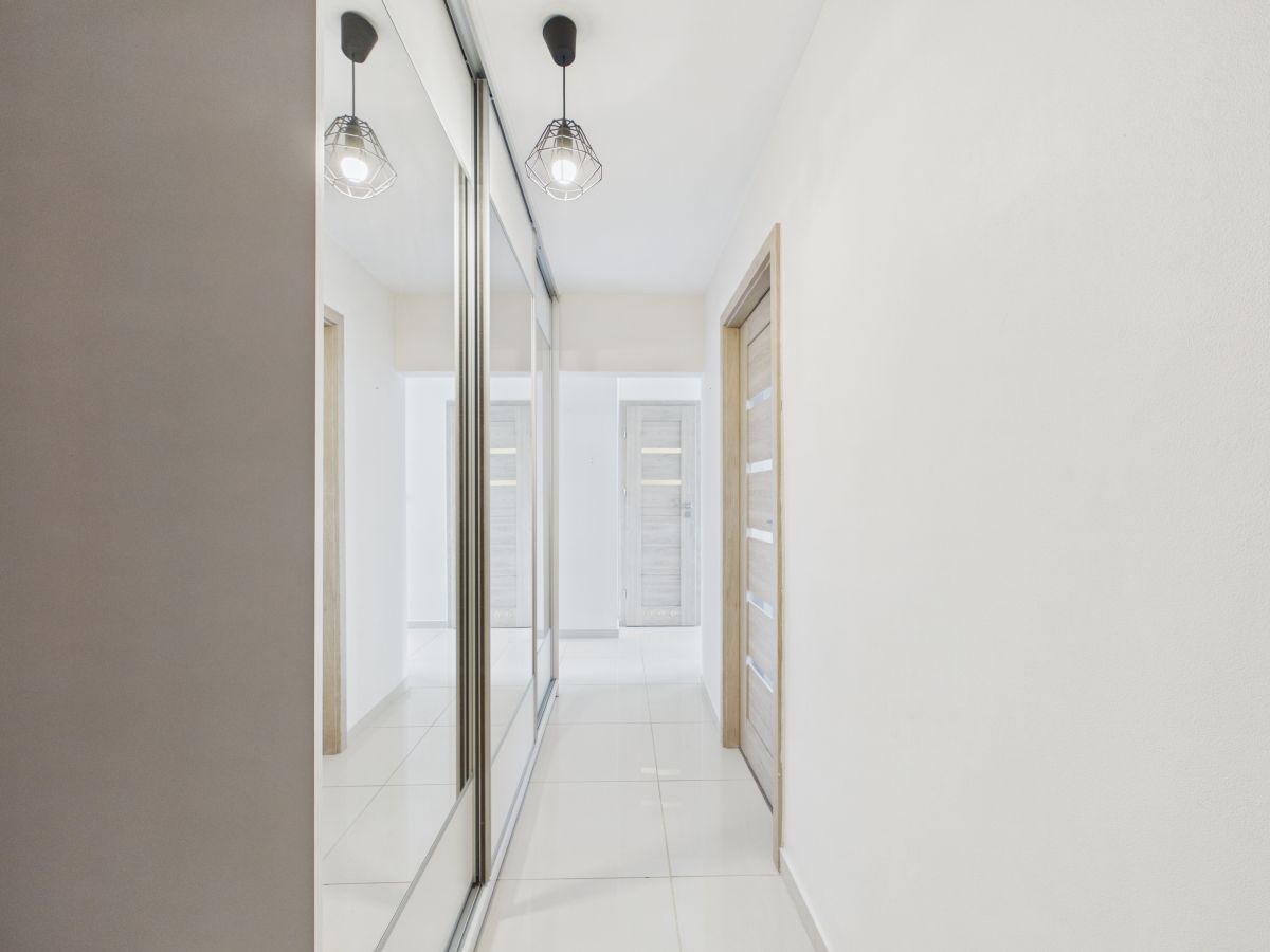 Bright, minimalist hallway with glossy white tile floor, mirrored closets on the left, and light wood doors on the right.