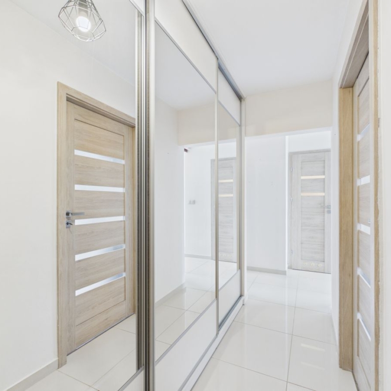 Bright hallway with mirrored closet doors reflecting light, wooden interior doors, and glossy white tile floor in a modern home.