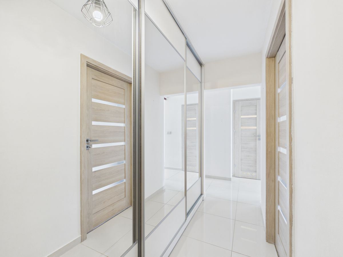 Bright hallway with mirrored closet doors reflecting light, wooden interior doors, and glossy white tile floor in a modern home.