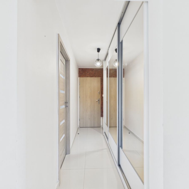 Narrow entry hallway with white walls, a wooden door at the end, brick accent wall, and a large mirrored wardrobe on the right.