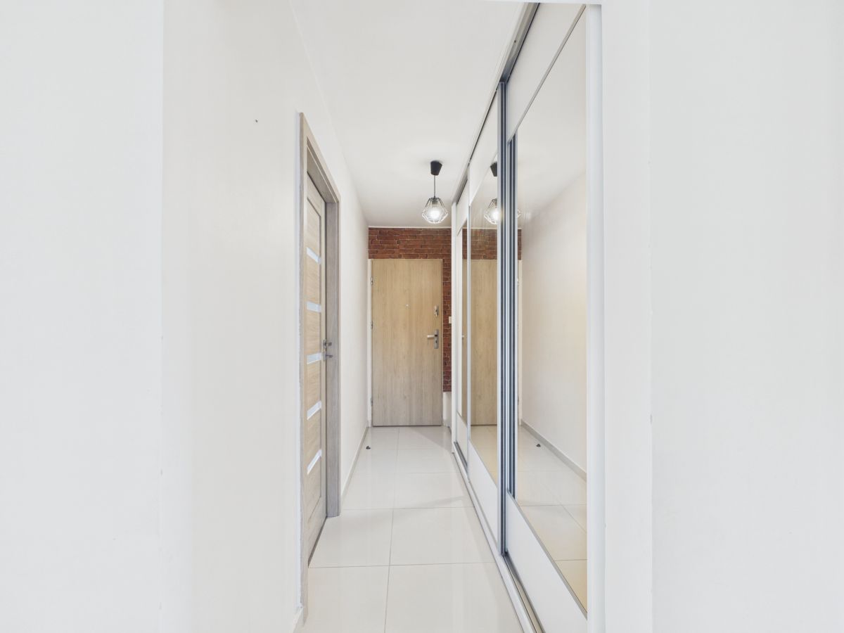 Narrow entry hallway with white walls, a wooden door at the end, brick accent wall, and a large mirrored wardrobe on the right.