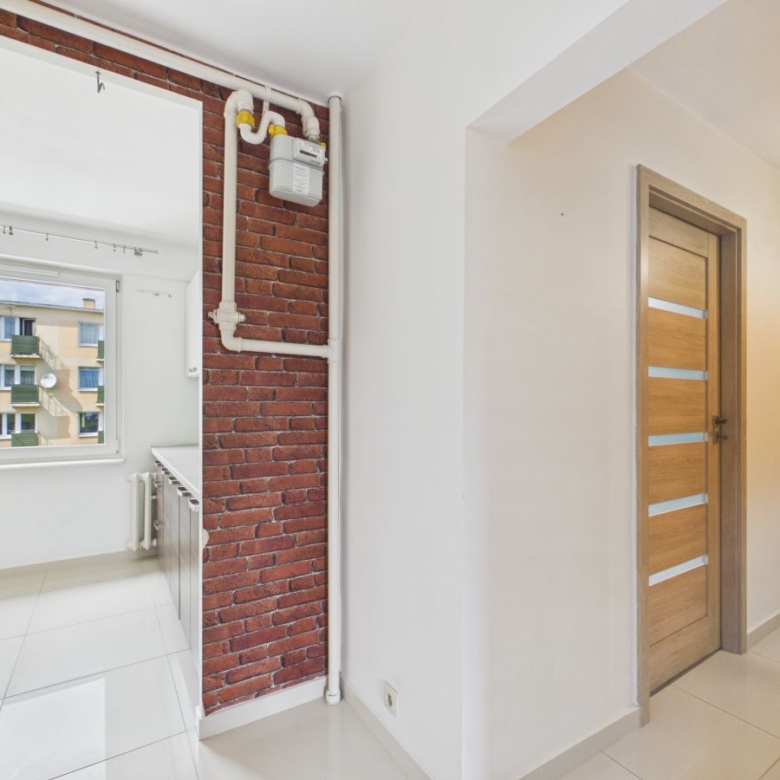 Bright hallway with white walls, a brick partial wall with exposed pipes and a gas meter, and a wooden door with frosted glass panels to the right.
