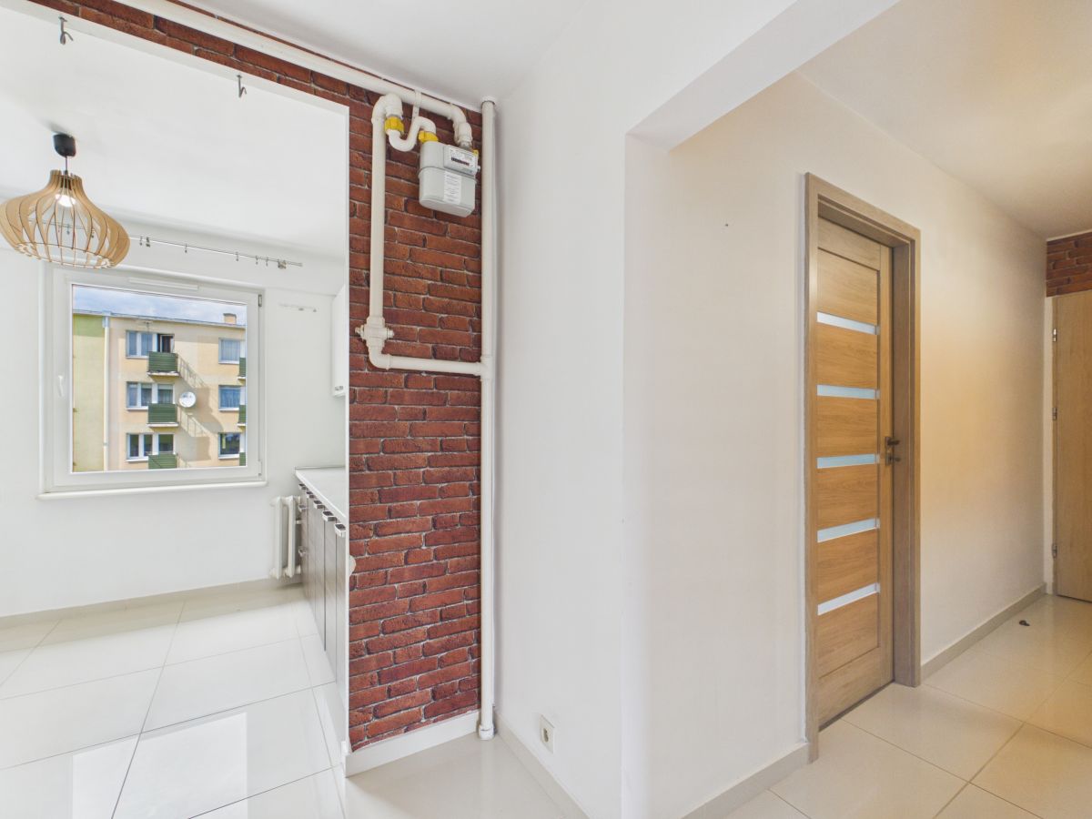 Bright hallway with white walls, a brick partial wall with exposed pipes and a gas meter, and a wooden door with frosted glass panels to the right.