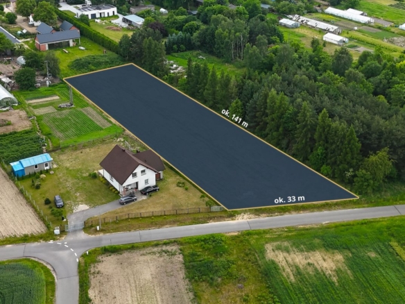 Aerial view of a rectangular plot outlined in yellow beside a white house, dimensions ~141 m by 33 m, in a rural area with fields and trees nearby.