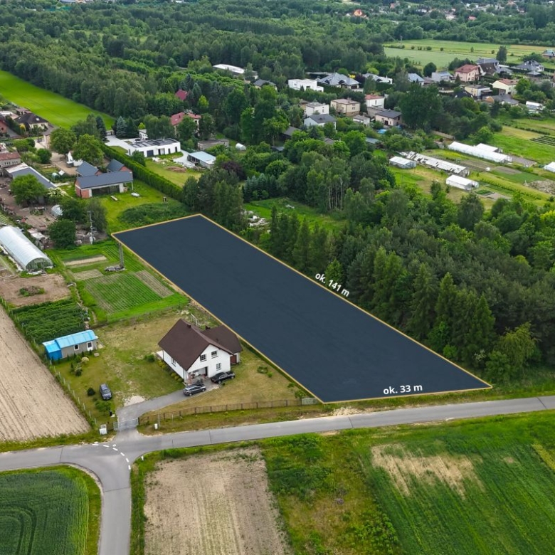 Rectangular vacant lot along a rural road, outlined for development, about 141 m by 33 m, surrounded by trees, gardens, and scattered houses in a countryside setting.