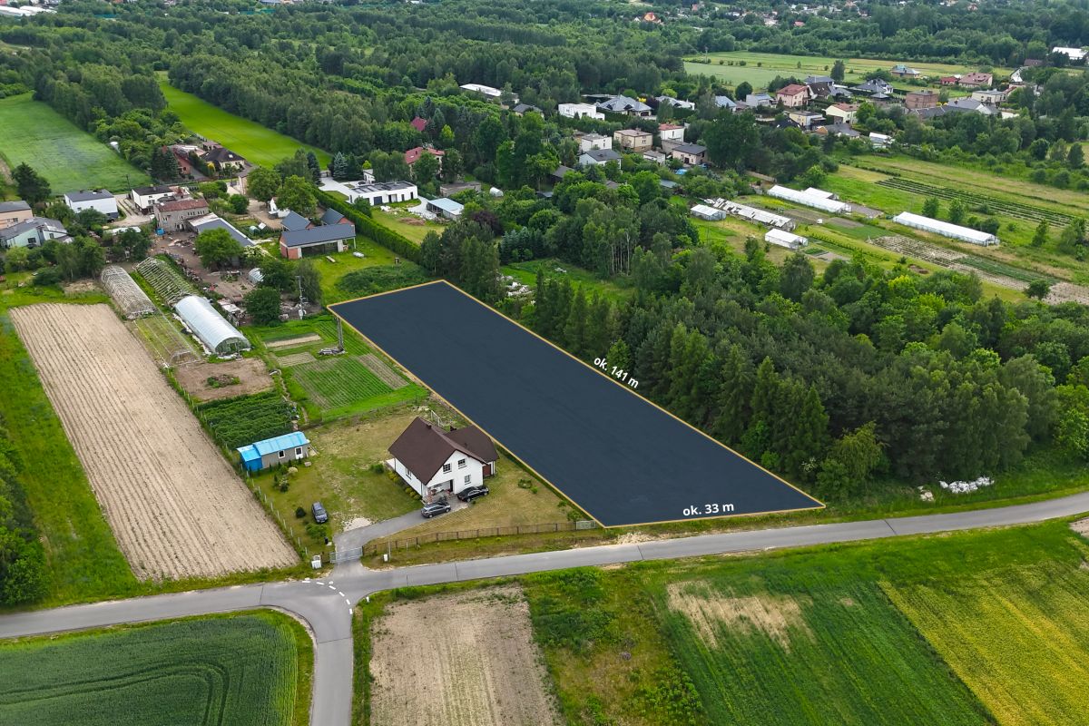 Rectangular vacant lot along a rural road, outlined for development, about 141 m by 33 m, surrounded by trees, gardens, and scattered houses in a countryside setting.