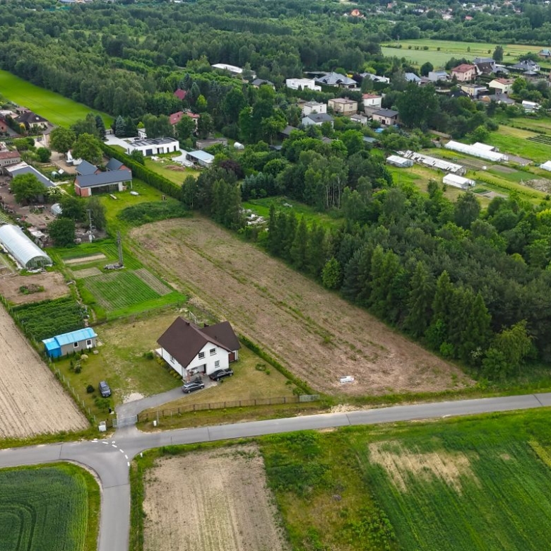 Aerial view of a rural village with fields, greenhouses, and a white house by a road intersection surrounded by trees and farmland