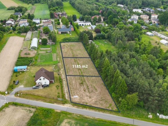 Aerial view of a residential area with a fenced, outlined vacant plot labeled 1185 m2 along a road, surrounded by trees and neighboring houses.