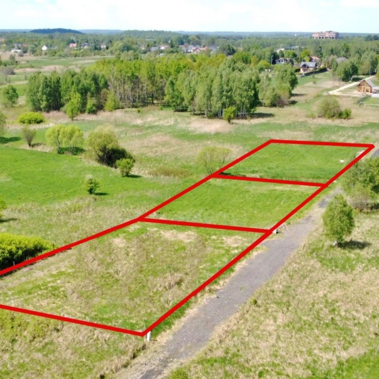 Aerial view of a grassy field with three rectangular land plots outlined in red along a rural road, with trees and distant houses in the background.