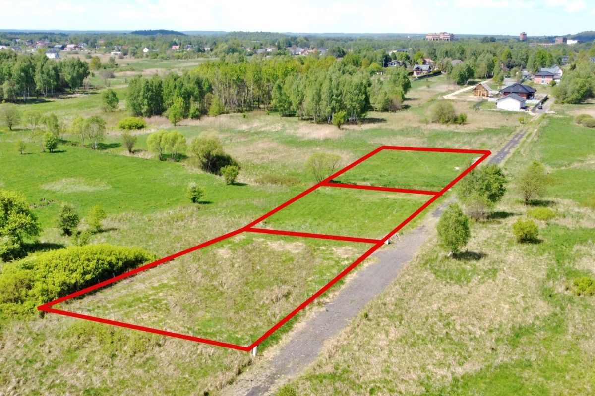 Aerial view of a grassy field with three rectangular land plots outlined in red along a rural road, with trees and distant houses in the background.