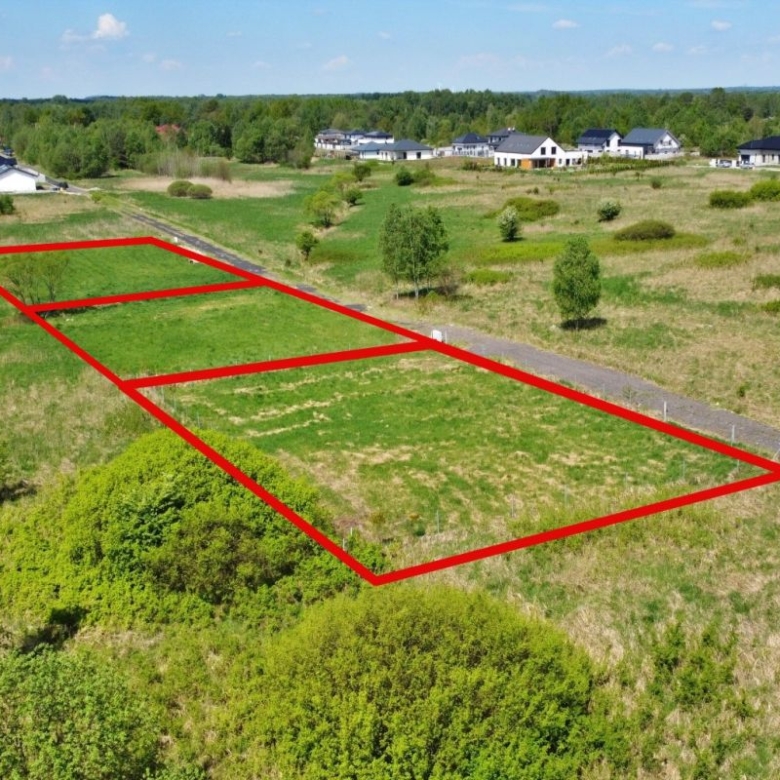 Aerial view of three building plots outlined in red along a road in a developing neighborhood on grassy land near houses in the distance (for sale).
