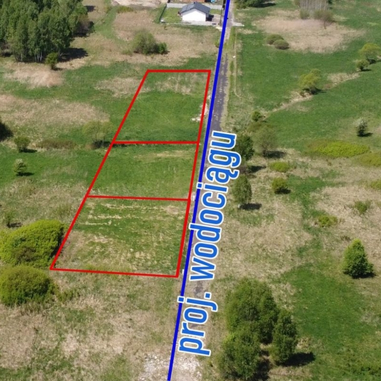 Aerial view of three red-outlined rectangular land plots next to a blue line labeled with text, in a grassy rural field with trees nearby.