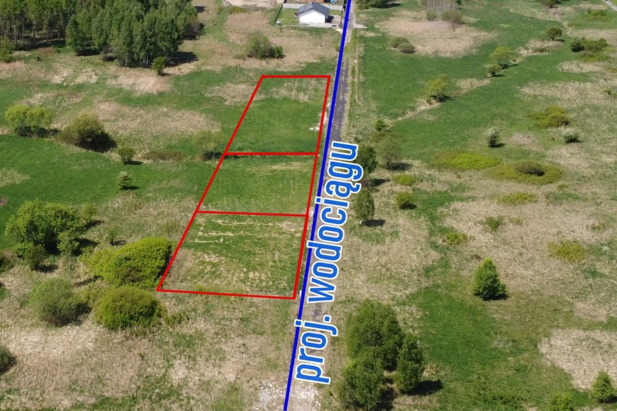 Aerial view of three red-outlined rectangular land plots next to a blue line labeled with text, in a grassy rural field with trees nearby.