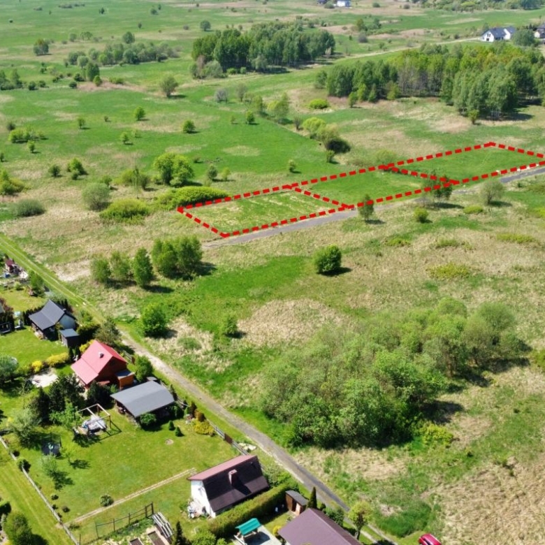 Aerial view of two adjacent vacant lots outlined in red beside a road in a rural area, with green fields and houses nearby.
